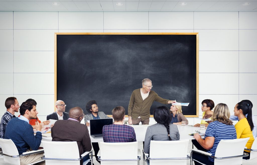 A gray haired man stands in front of a large blackboard and addresses a multiethnic group of students. The students are sitting around a large table.